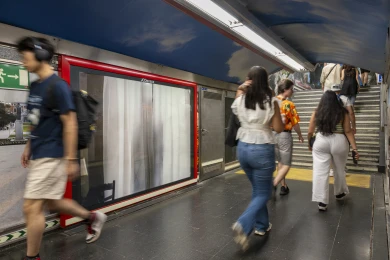 La misma valla en el andén del metro, con viajeros caminando hacia las escaleras
