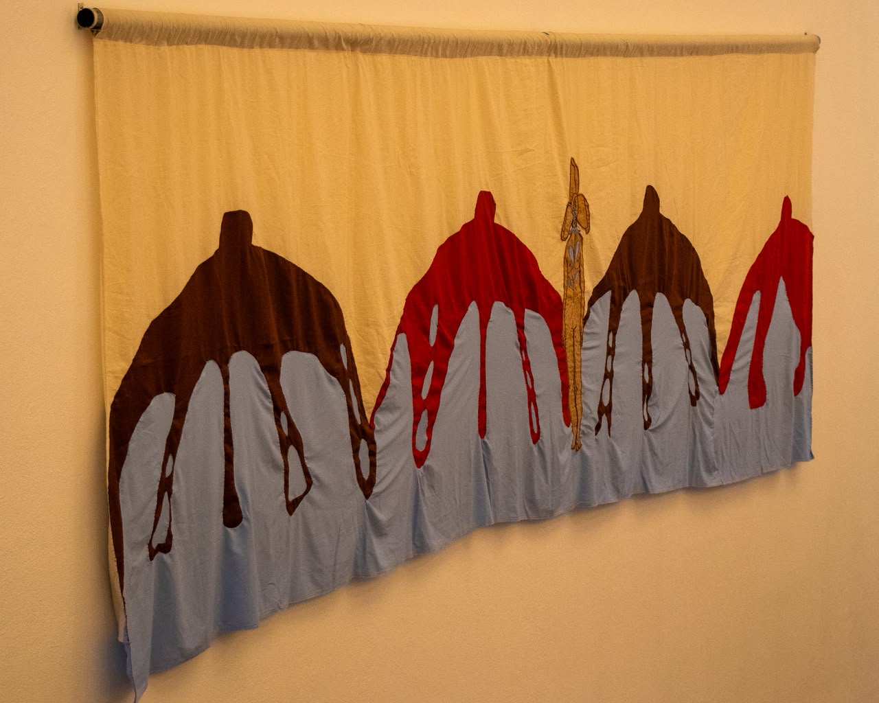 Gran textil cosido, con siluetas rojas, marrones y celestes, colgado de una barra en la sala.